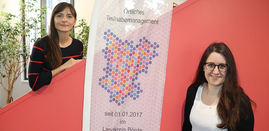 Das Foto zeigt die Innentreppe des Landratsamtes in Haldensleben, davor ein Banner mit der Aufschrift "Örtliches Teiilhabemanagement" und Katja Klommhaus (links) und Anne Sophie Fischer als direkte Ansprechpersonen für Angelegenheiten von Menschen mit Beeinträchtigungen. 