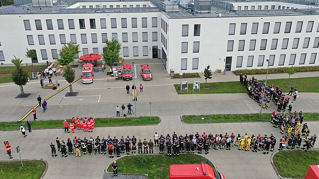 Die Luftbildaufnahme ist am 11. September 2021 zum Übungsbeginn mit einer Drohne am Verwaltungsgebäude des Landkreises in Haldensleben entstanden. Es zeigt die an der Übung beteiligten Einheiten, Menschen und Technik, aus der Vogelperspektive. Im Vordergrund haben die Einheiten Aufstellung auf dem Parkplatz genommen. Im Hintergrund ist das Verwaltungsgebäude "Bornsche Straße 2" zu sehen.