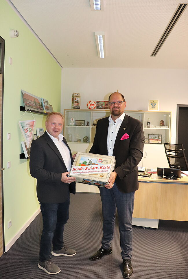 Foto Landkreis Börde / Landrat Steve Kanitz auf Stippvisite bei seinem Amtskollegen im Landkreis Börde. Beide Herren stehen im Büro von Martin Stichnoth nebeneinander. Stichnoth überreicht eine "Börde Schatz Kiste".