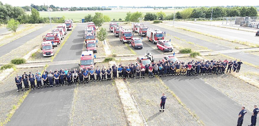 Foto / Landkreis Börde / die Helfer haben sich am Nachmittag des 21. Juni 2022 in Hersdorf gesammelt, um gemeinsam den Marsch in das Waldbrandgebiet nach Beelitz anzutreten. Hier ein Luftbild auf die Fahrzeuge und die Einsatzkräfte. Sie erhalten den Marschbefehl von Kreisbrandmeister Matthias Schumann.