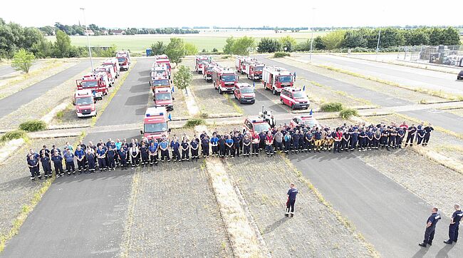 Foto / Landkreis Börde / die Helfer haben sich am Nachmittag des 21. Juni 2022 in Hersdorf gesammelt, um gemeinsam den Marsch in das Waldbrandgebiet nach Beelitz anzutreten. Hier ein Luftbild auf die Fahrzeuge und die Einsatzkräfte. Sie erhalten den Marschbefehl von Kreisbrandmeister Matthias Schumann.