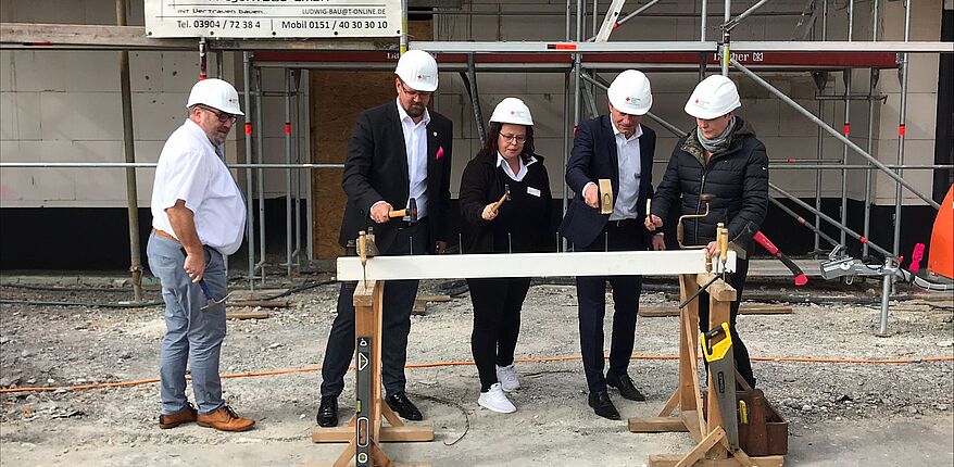 Das Foto zeigt im Hintergrund einen kleinen Ausschnitt des Rohbaugebäudes. Davor ein Holzbock, in den Ralf Kürbis und Sylvia Franke (beide DRK), die Pflegedienstleiterin des Wohnquartiers Oebisfelde, Katja Meinke, und der Landrat die letzten Nägel vor dem Richtfest eintreiben. Beobachter des Geschehens ist Hans-Werner Kraul, Bürgermeister von Oebisfelde-Weferlingen.   