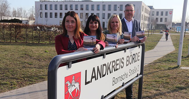 Stefanie Kunz, Netzwerkkoordinatorin, Familienhebamme und Koordinatorin Christina Stolarczyk präsentieren den Beratungswegweiser. Sie stehen hinter dem Einfahrtsschild mit der Aufschrift "Landkreis Börde" zum Parkplatz des Verwaltungsgebäudes in Haldensleben.