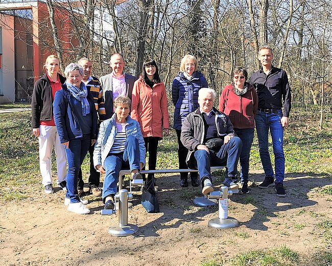 Foto Stadt Haldensleben / Sabine Wendler, Rüdiger Mages und Dr. Elena Sterdt freuen sich mit den Gästen über die Einweihung des Bewegungs-Parcours in Althaldensleben. Im Vordergung wird die Beinpresse getestet. Im Hintergrund stehen die gäste der Einweihung zum Erinnerungsfoto. 