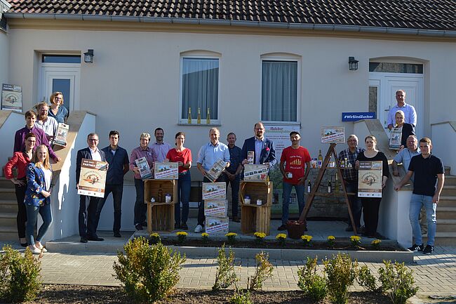 Das Foto mit 20 Personen, darunter Landrat Martin Stichnoth, ist auf dem Hof von Obstbauer Marcus Nickel in Hötensleben entstanden. Auf 2 Teppen zeigen die Direktvermarkter vor einem Gebäude die "Börde Schatz Kiste" und darauf hinweisende Werbematerialien