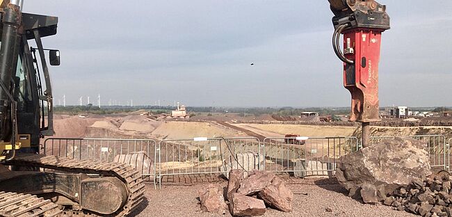 Das Foto zeigt einen seitlichen Einblick in den Tagebau bei Mammendorf. Hier wird das Gestein des Jahres 2020 (Andesit) gewonnen. Im Vorgergrund ist ein Kettenfahrzeug mit einem schweren Industriemeißel an einem Gesteinsblock zu sehen.