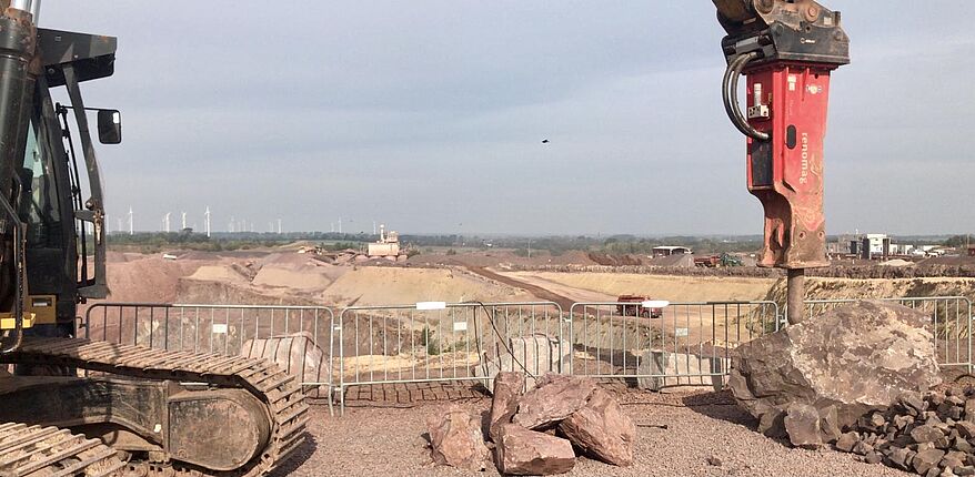 Das Foto zeigt einen seitlichen Einblick in den Tagebau bei Mammendorf. Hier wird das Gestein des Jahres 2020 (Andesit) gewonnen. Im Vorgergrund ist ein Kettenfahrzeug mit einem schweren Industriemeißel an einem Gesteinsblock zu sehen.