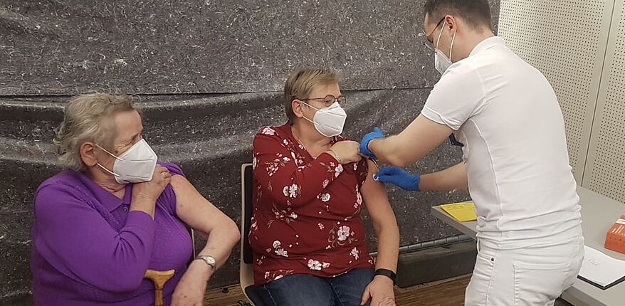 Das Foto zeigt einen impfberechtigte Person bei der Impfung. Eine Person wird gerade geimpft, eine zweite Person wartet. 